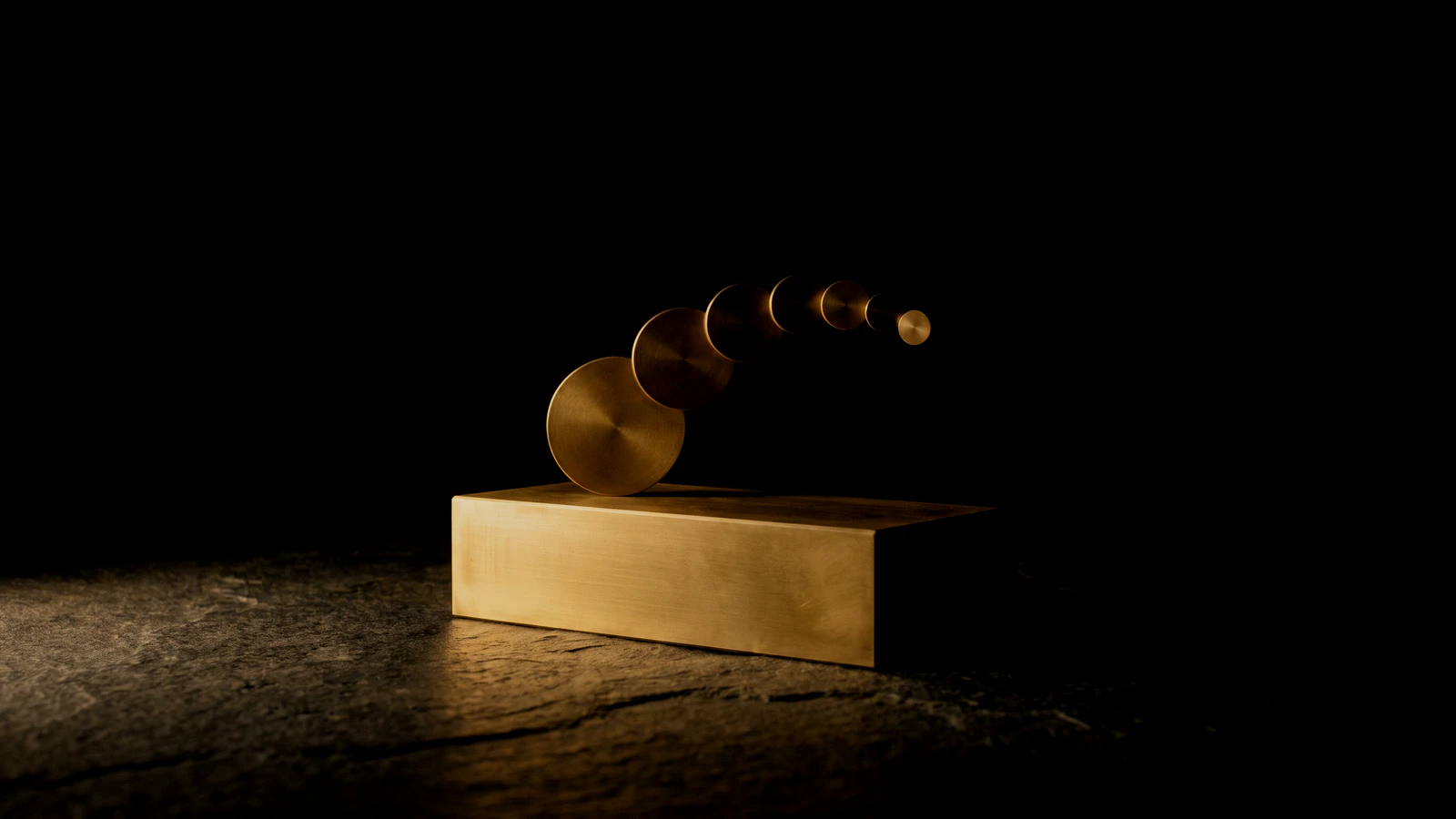 A heavy brass block on black with smaller brass discs stepping upward away from it.